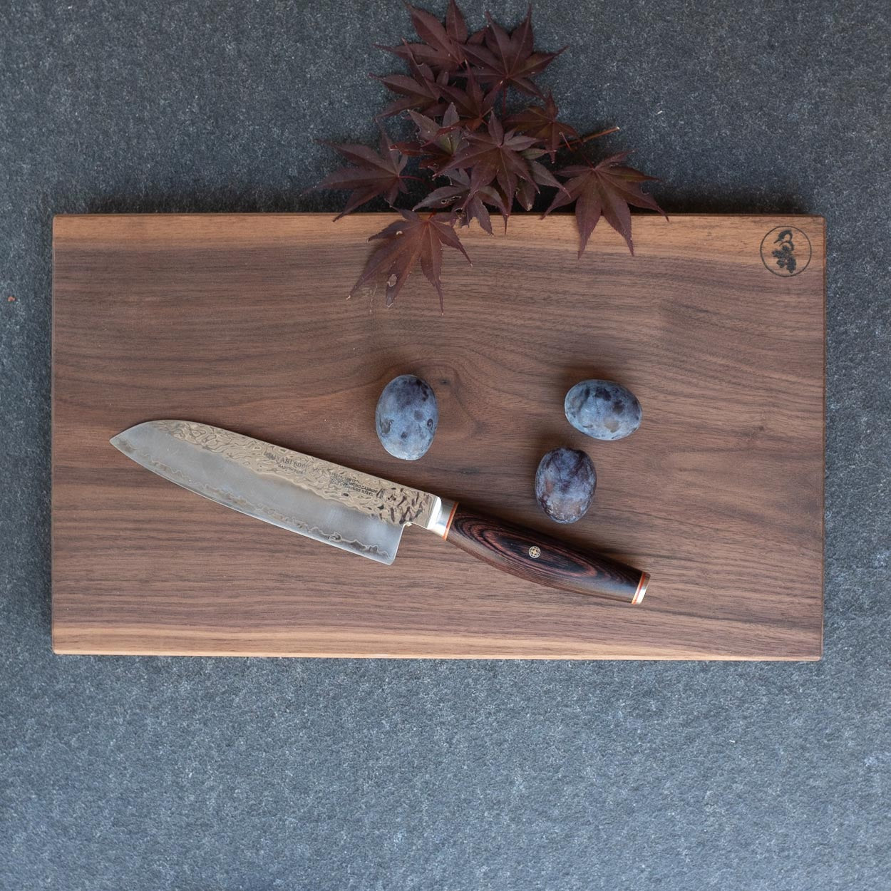 Nussbaum Schneidebrett von oben mit einem Santoku Messer und drei Pflaumen.
