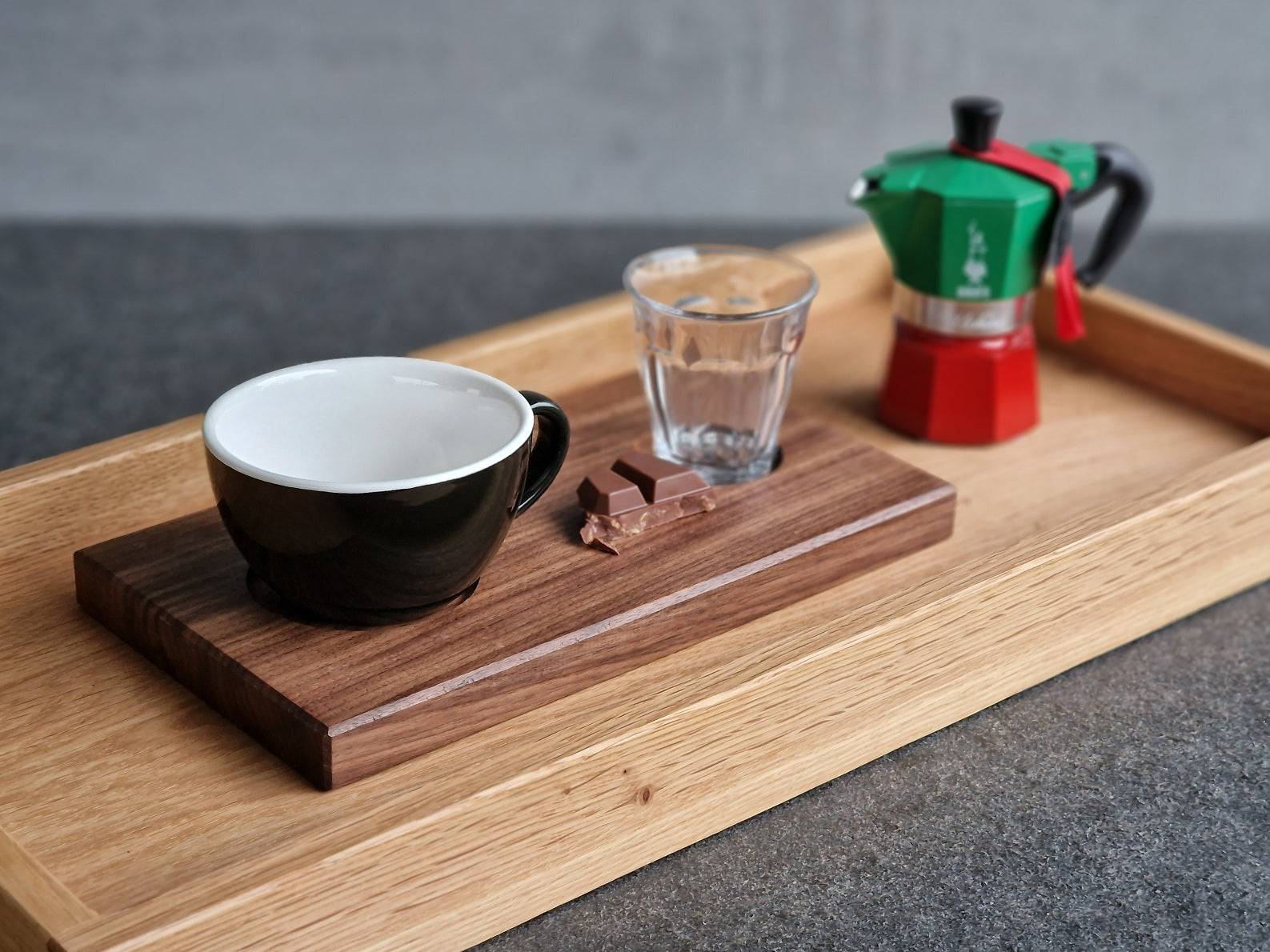 Auf einem Eiche Holztablett steht ein kleines Nussbaum Tablett mit einer Cappuccinotasse und einem Wasserglas. Daneben eine Bialetti Moka Express.