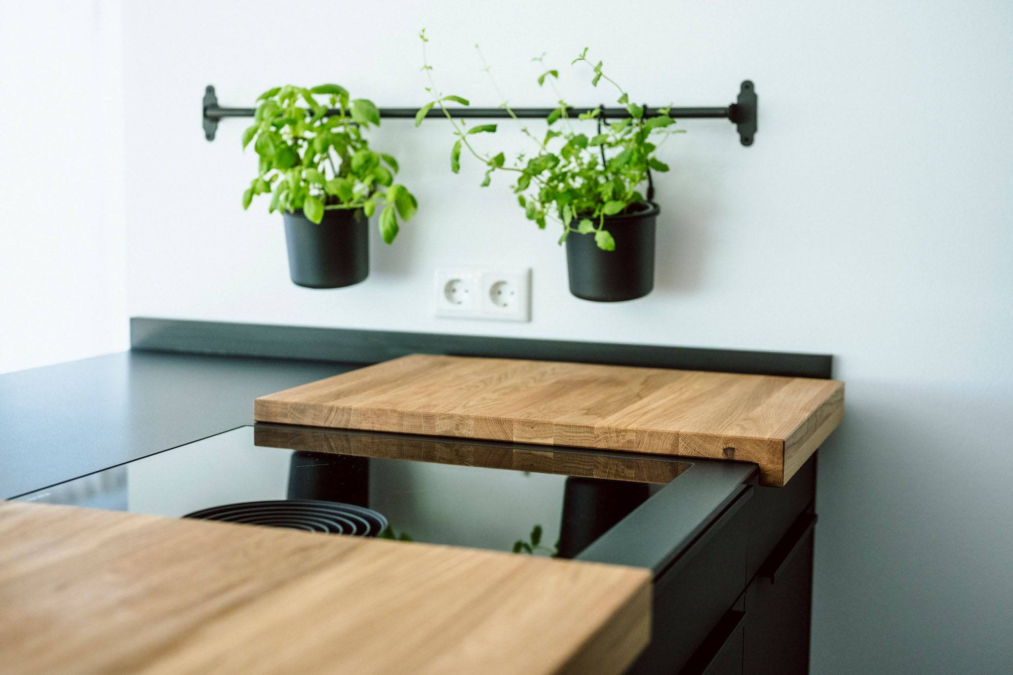 Zweiteilige Kochfeldabdeckung auf einem Bora Professional Kochfeld, das Kochfeld ist auf einer schwarzen Design Kücheninsek montiert mit schwarzen Fronten. An der weißen Wand ist eine Blumenleiste an der zwei Eimer mit Gartenkräuter hängen