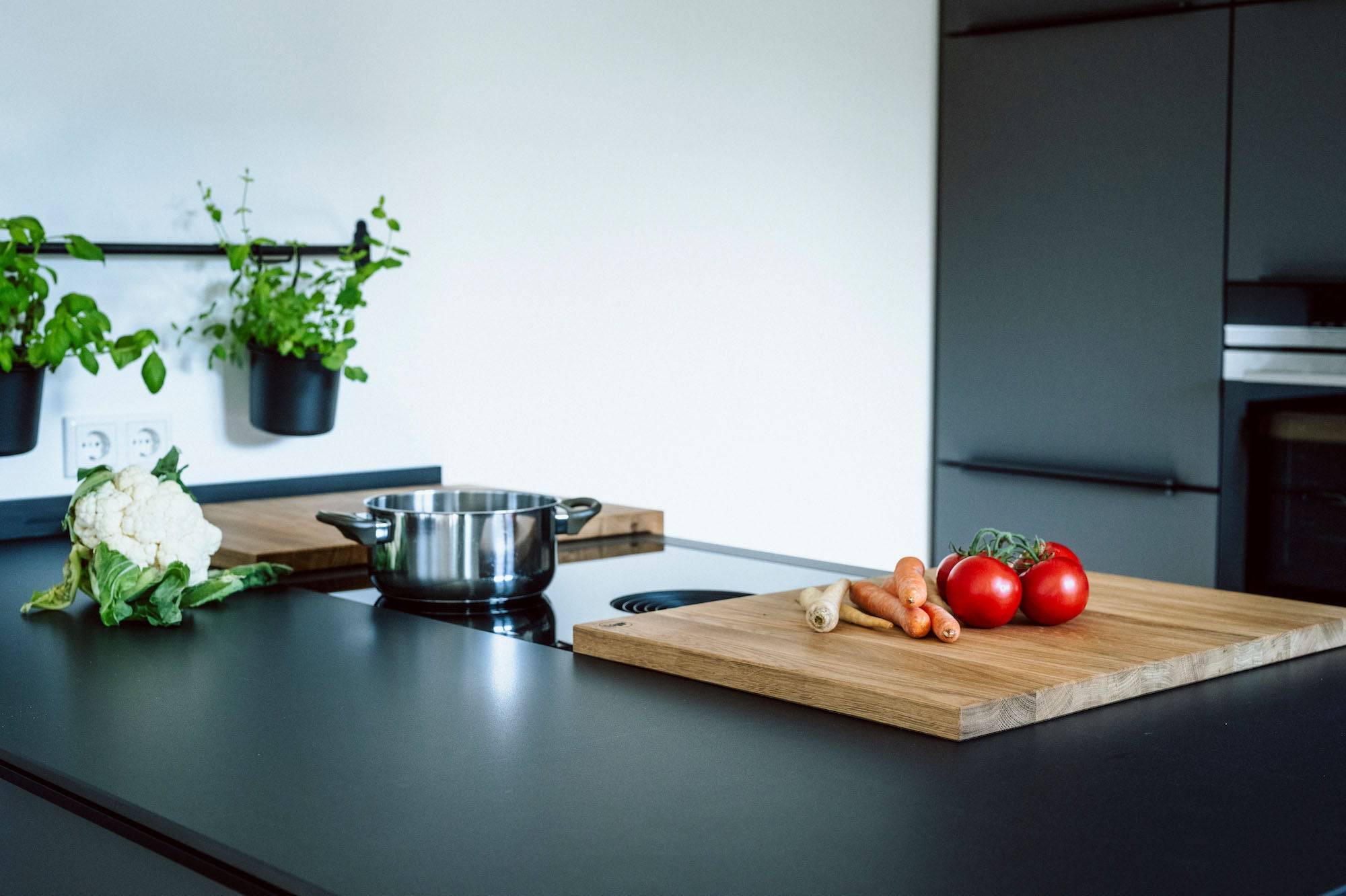 Schwarze Kücheninsel mit Bora Kochfeld auf dem ein Eichen Kochfeldschutz liegt. Auf dem Kochfeld steht noch eine Edelstahltop auf der Kücheninsel liegt ein frischer Blumenkohl. Eine weiße Wand im Hintergrund.