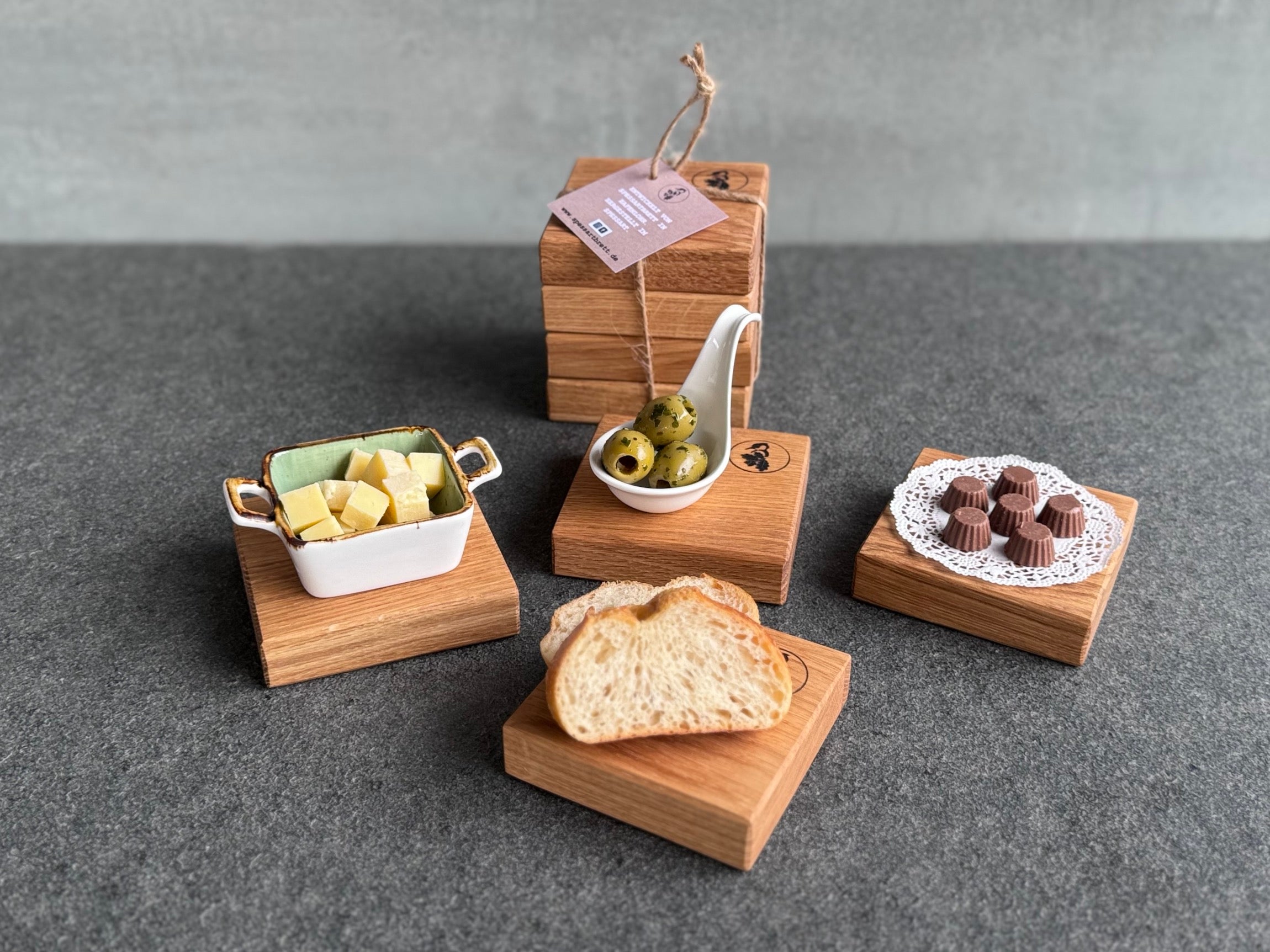 Als Geschenk verschnürte Eichen Untersetzer. Im Vordergrund 4 kleine Eichen Servierbrettchen mit Käse, Oliven, Pralinen und Baguette.