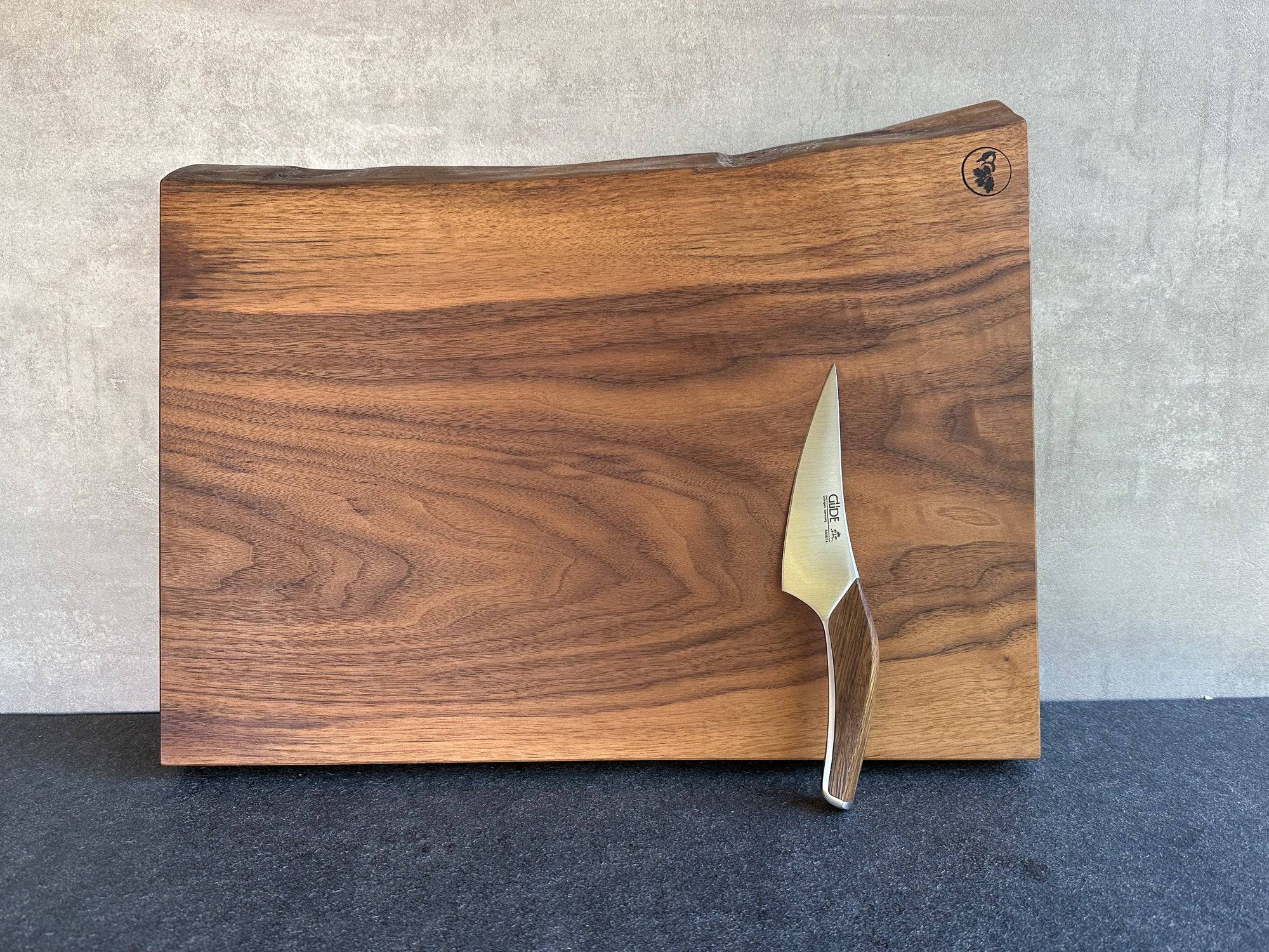 Aus einem Stück Nussbaum Vollholz: einzigartiger Schneideblock mit einer Baumkante und einem Güde Kochmesser dekoriert.