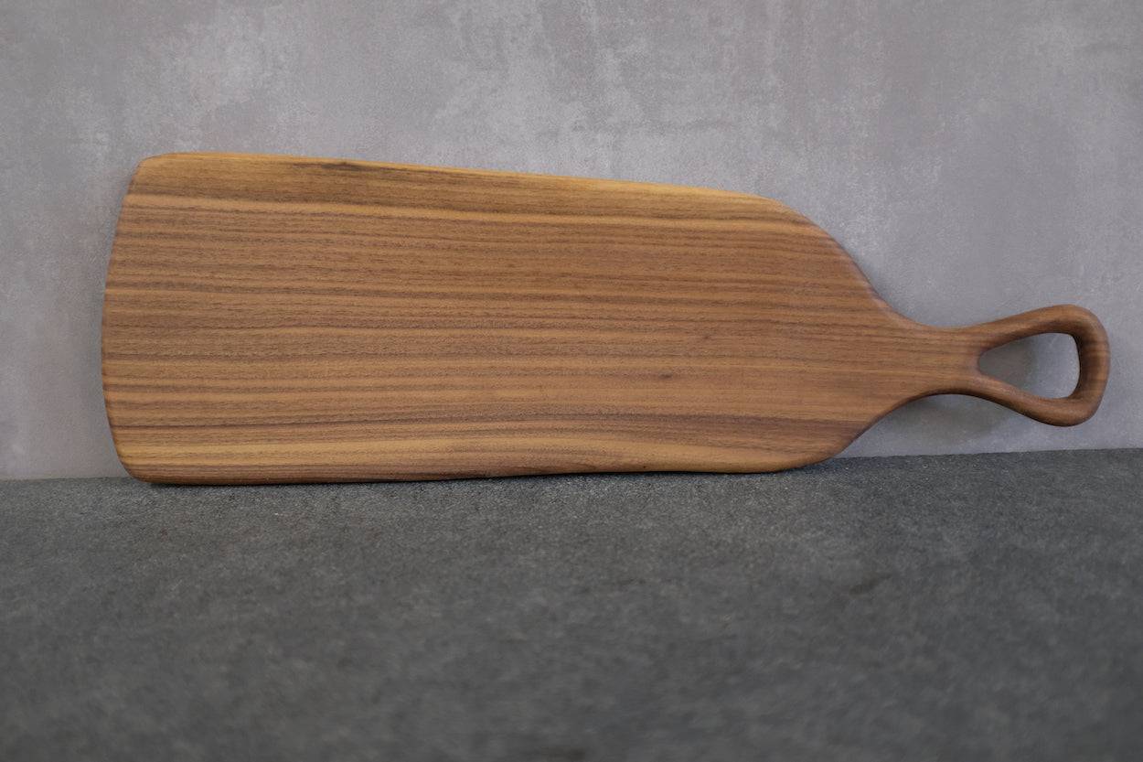 Dunkles Holz, massiv, aus einem Stück Nussbaum. Servierbrett mit Griff vor einer Betonwand.