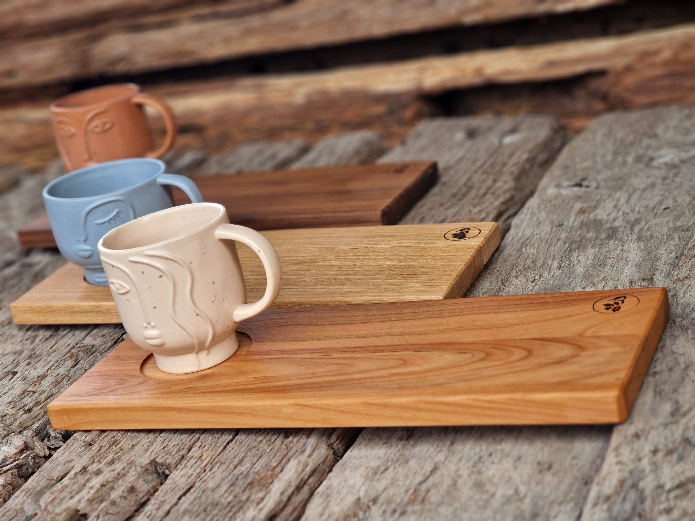 3 Holz Tabletts in Eiche, Kirsche und Nussbaum, jeweils mit einer Vertiefung für eine große Tasse. Dekoriert mit 3 Bechern mit Gesicht. Auf einem Eichenbalken.