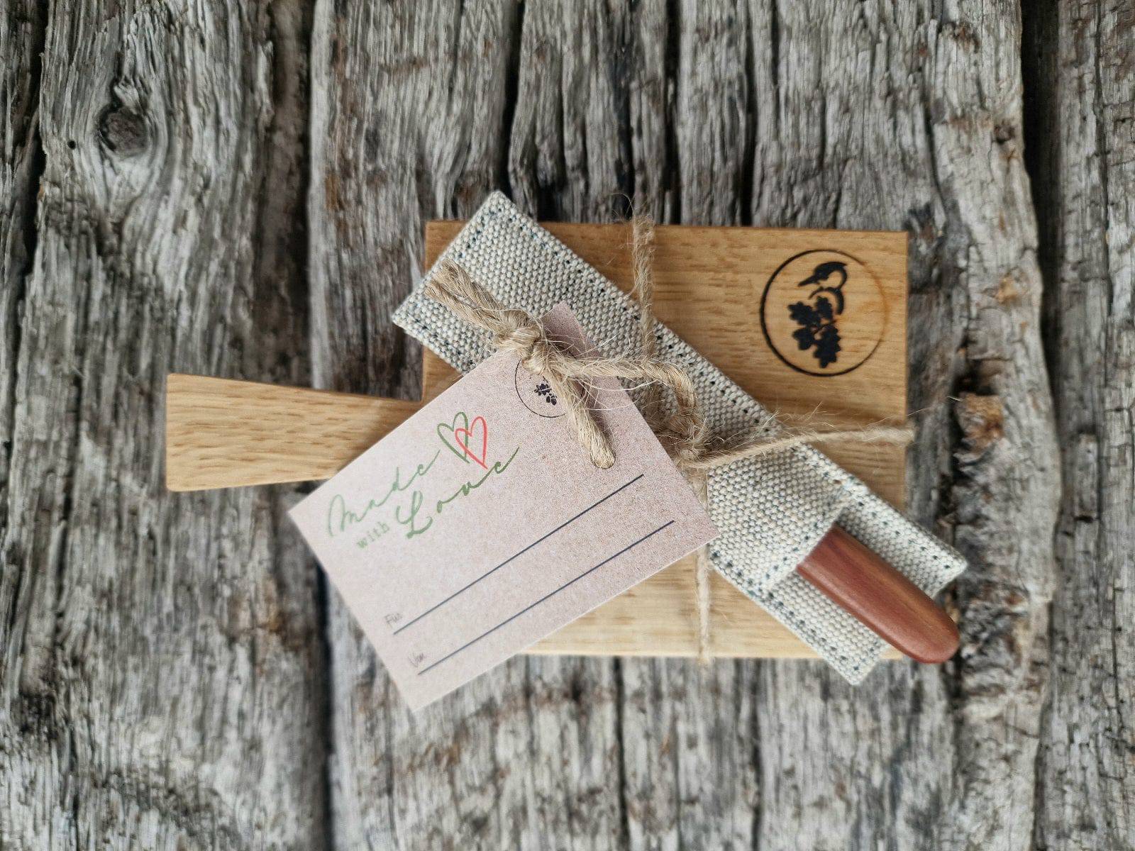 Eichenbrett für eine Baguette Scheibe, mit einem Buttermesser und einem passenden Etui aus festem Leinen. Fotografiert mit einem Geschenkanhänger auf alten Eichenbalken.