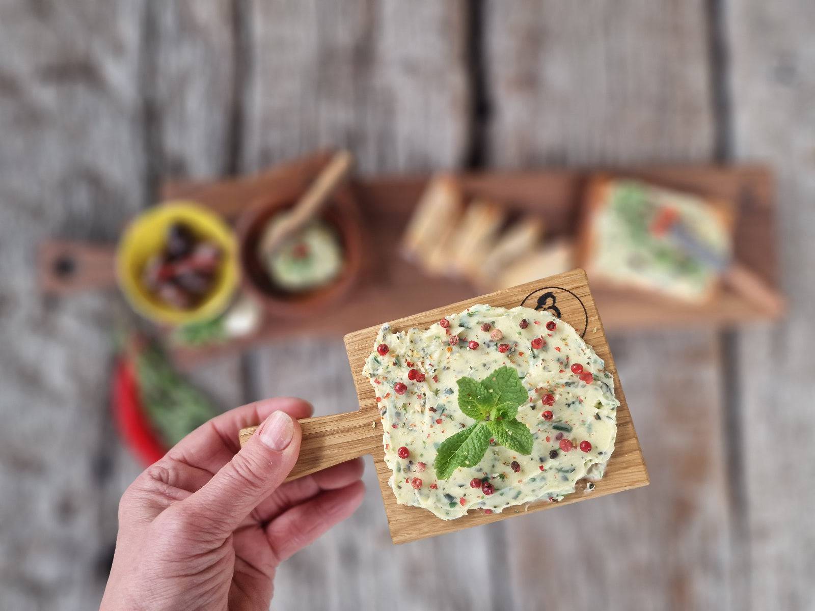 Ein Butterboard aus massiver Eiche in einer Hand. Im Hintergrund unscharf ein Servierbrett Aus Nussbaum mit verschieden Tapas dekoriert.