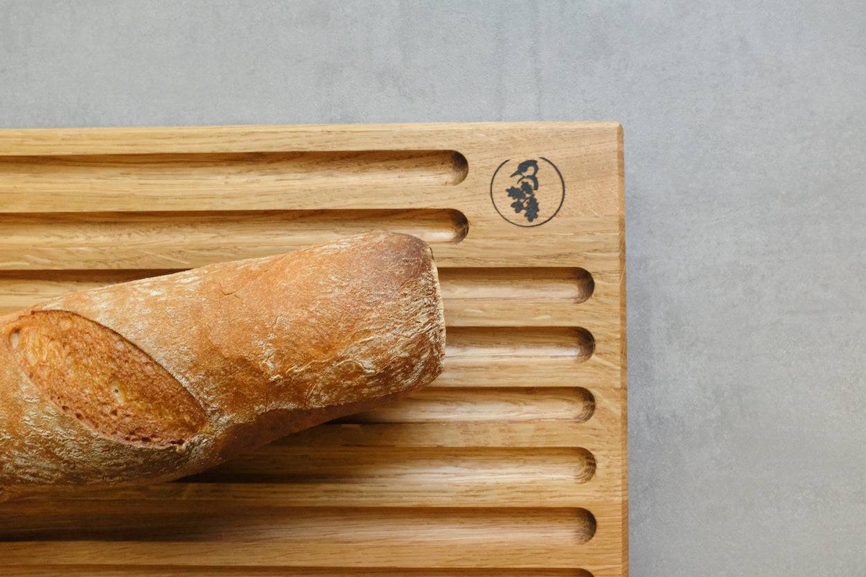 Detailfoto von einem Brotschneidebrett. Man sieht die rechte obere Ecke, 7 Rillen für Brösel. Auf dem Brotschneidebrett liegt ein Baguette
