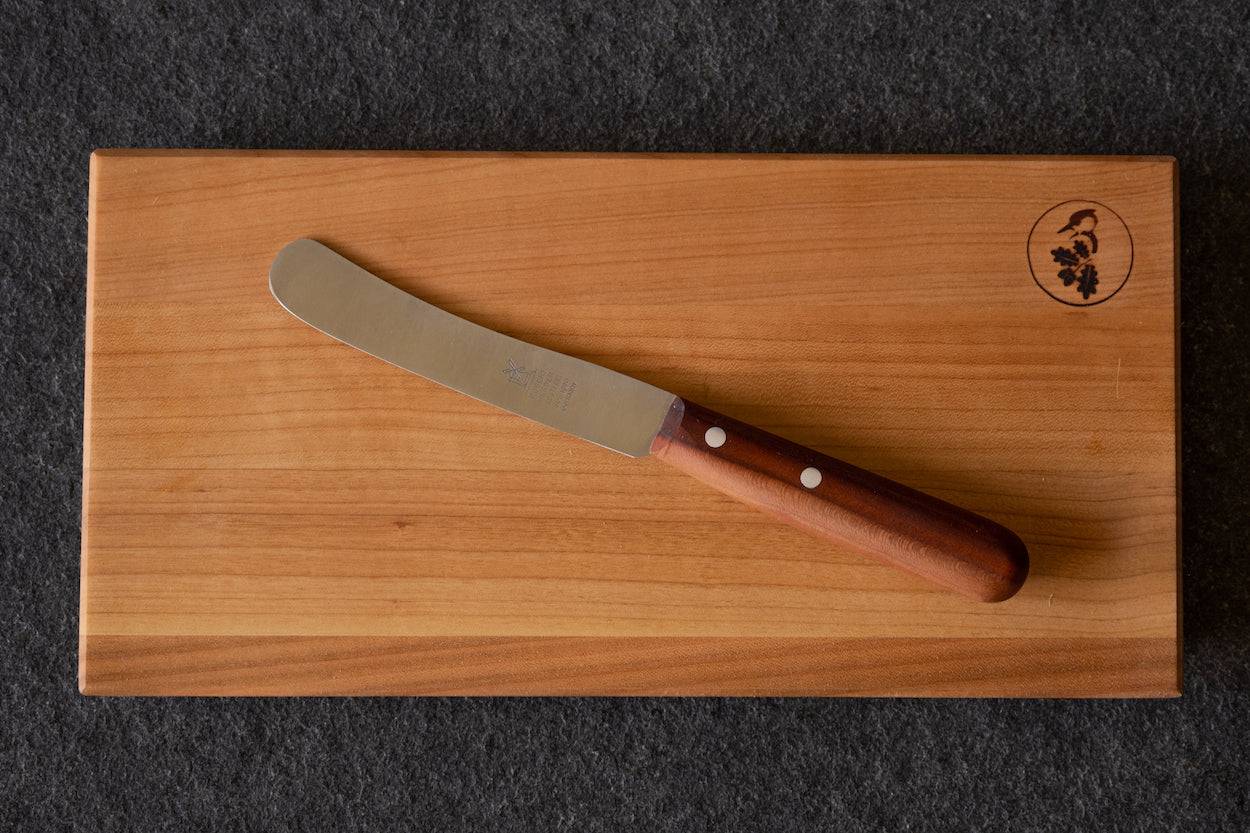 Buckels Tischmesser von Windmühlenmesser mit Pflaumengriff liegt auf einem Brett aus Kirschbaum.