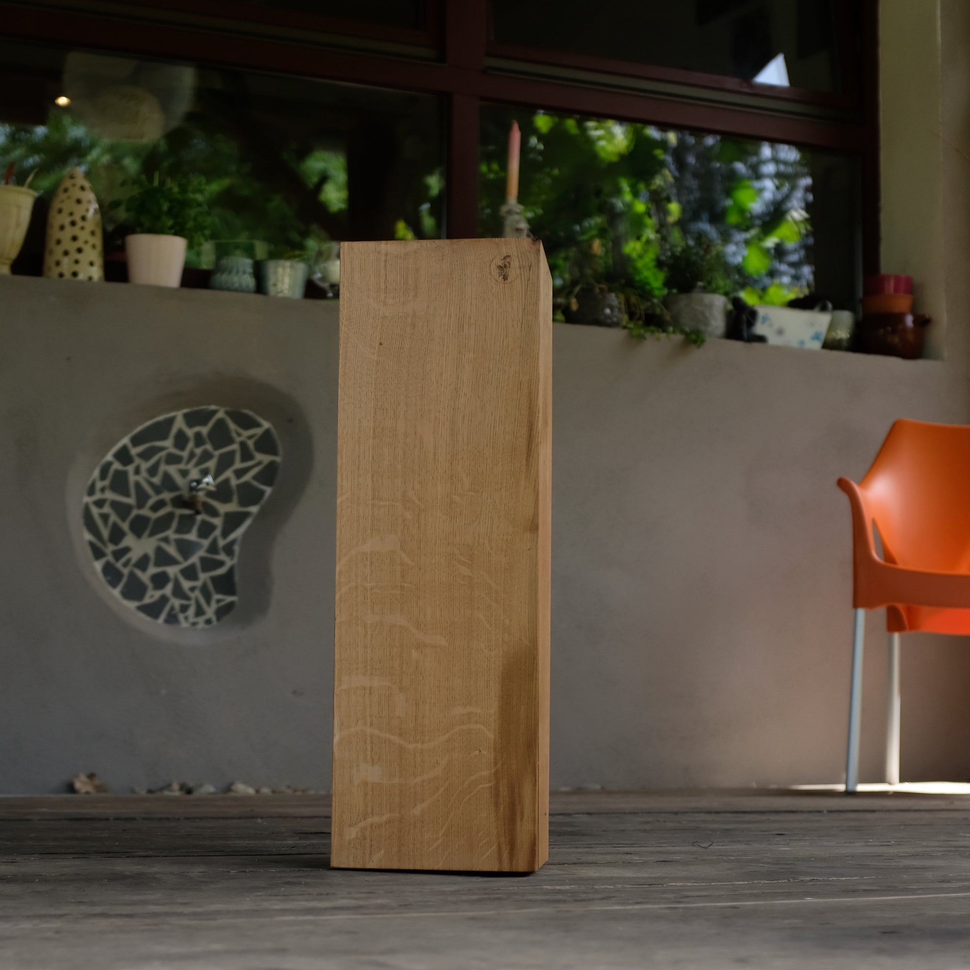 Eine Eichen Säule, die auf einer Terrasse steht. Im Hintergrund ein Wasserhahn an einer grauen Wand, ein Fenster, auf dem Blumen stehen und ein orangener Stuhl