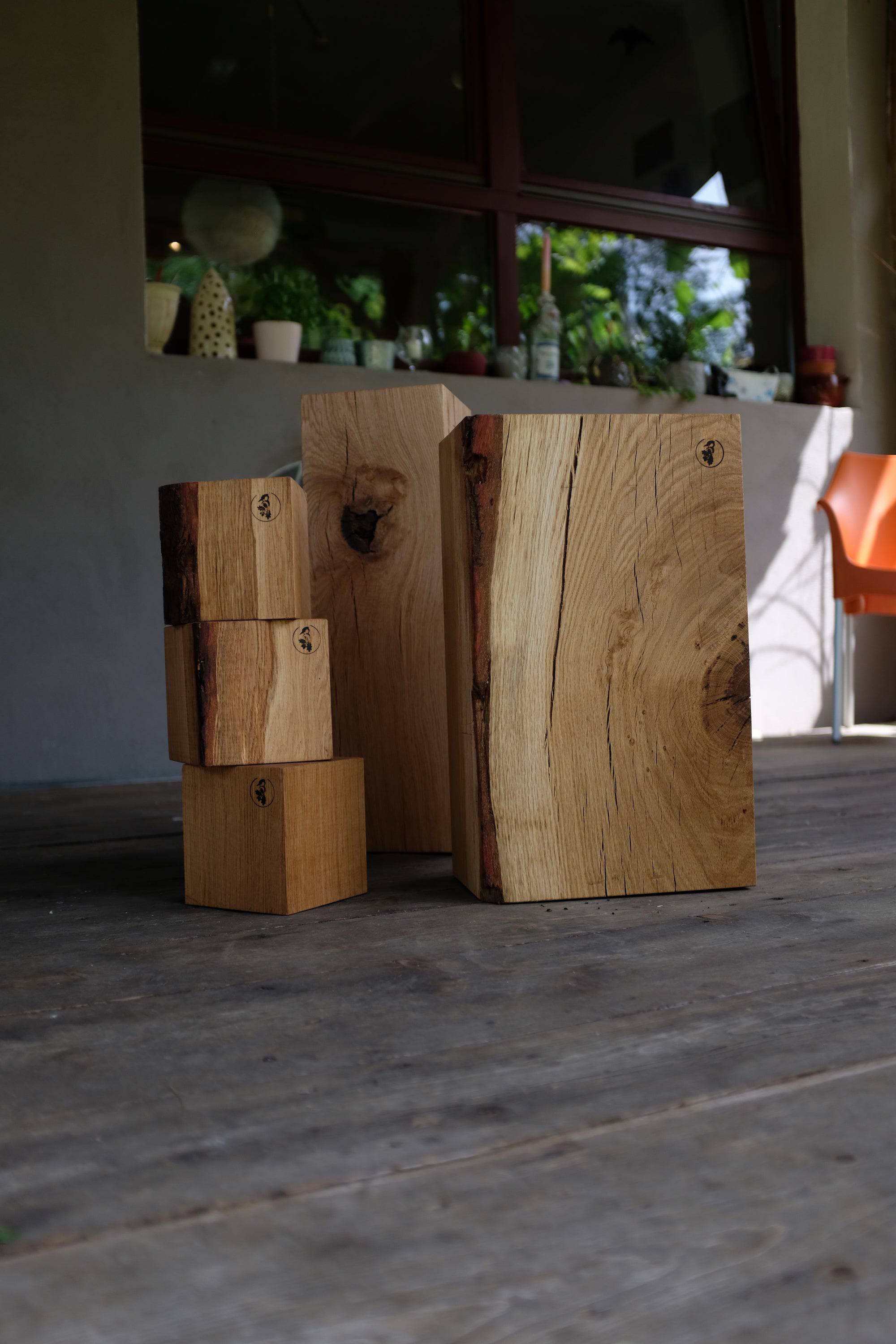 Drei Eichenklötze gestapelt und ein großer Eichenklotz, im Hintergrund ein Fenster mit Dekoration. Alles auf einem Terassenboden fotografiert