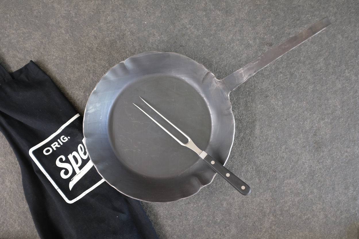 Eisenpfanne mit hohem Rand auf einer Steinplatte. Mit einer Fleischgabel und Spessart Kochschürze dekoriert.