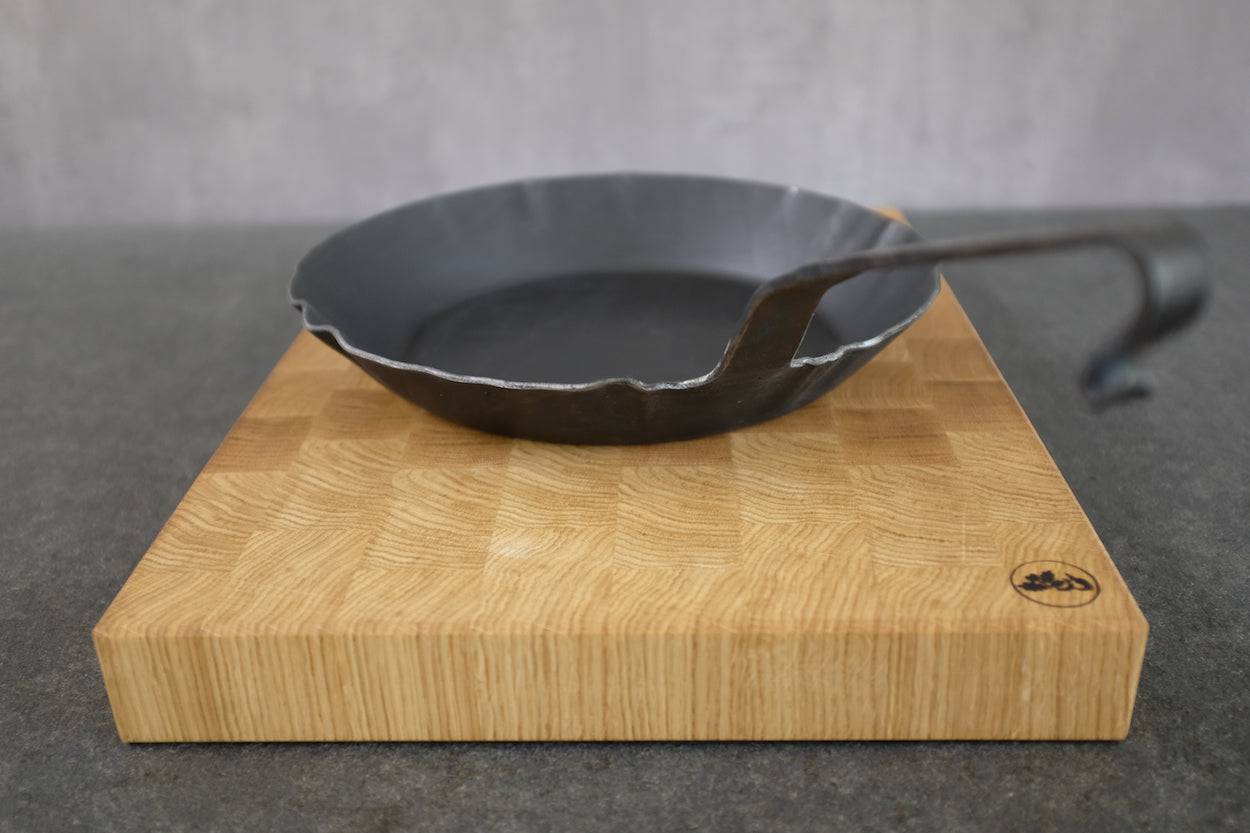 Geschwungener Hakenstiel einer handgeschmiedeten Eisenpfanne auf einem Eiche Stirnholz Schneidebrett.