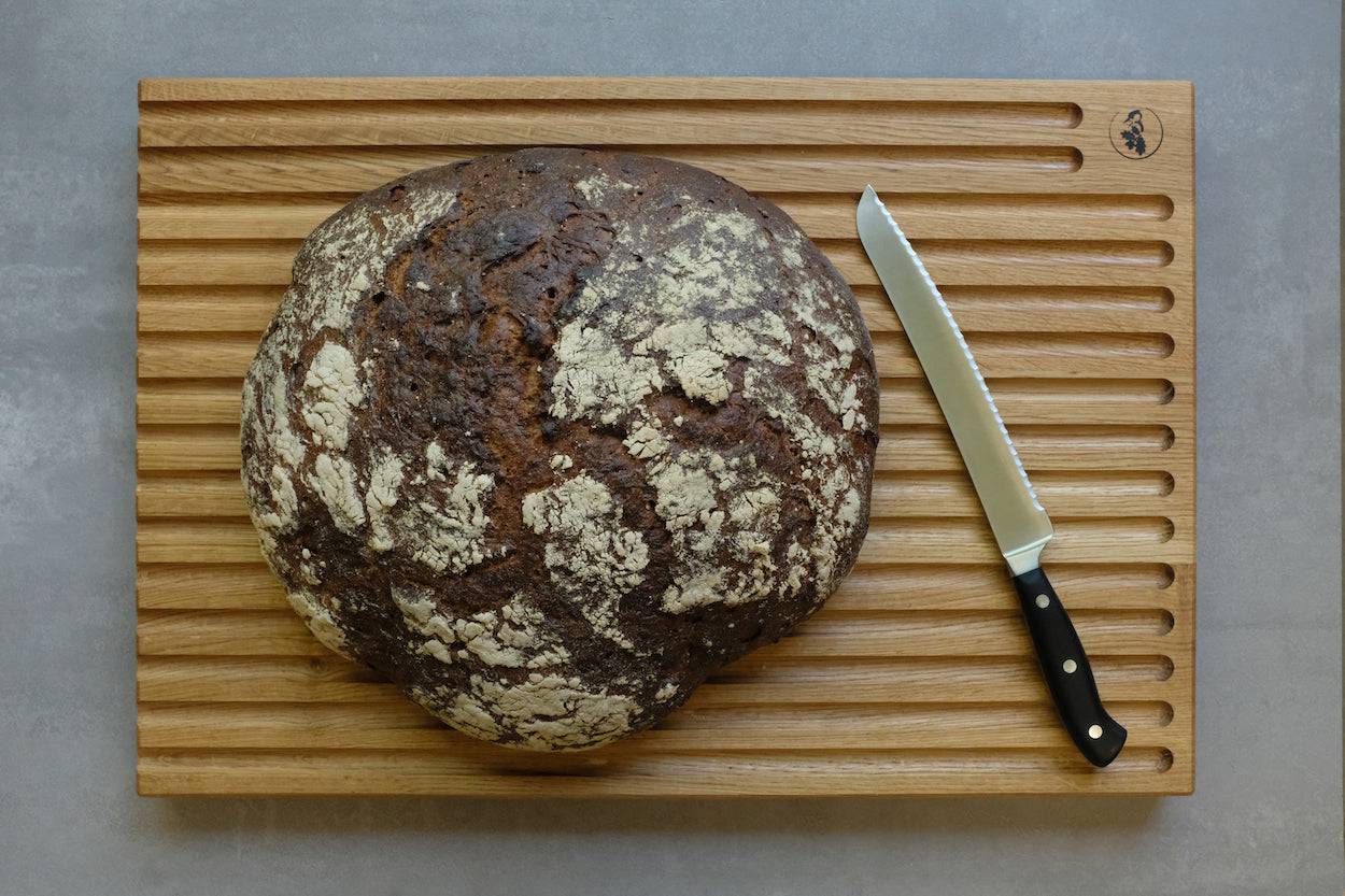 Ein Brotbrett mit Rillen. Auf dem Brett liegt ein großer Laib Brot und ein Brotmesser von Dick.