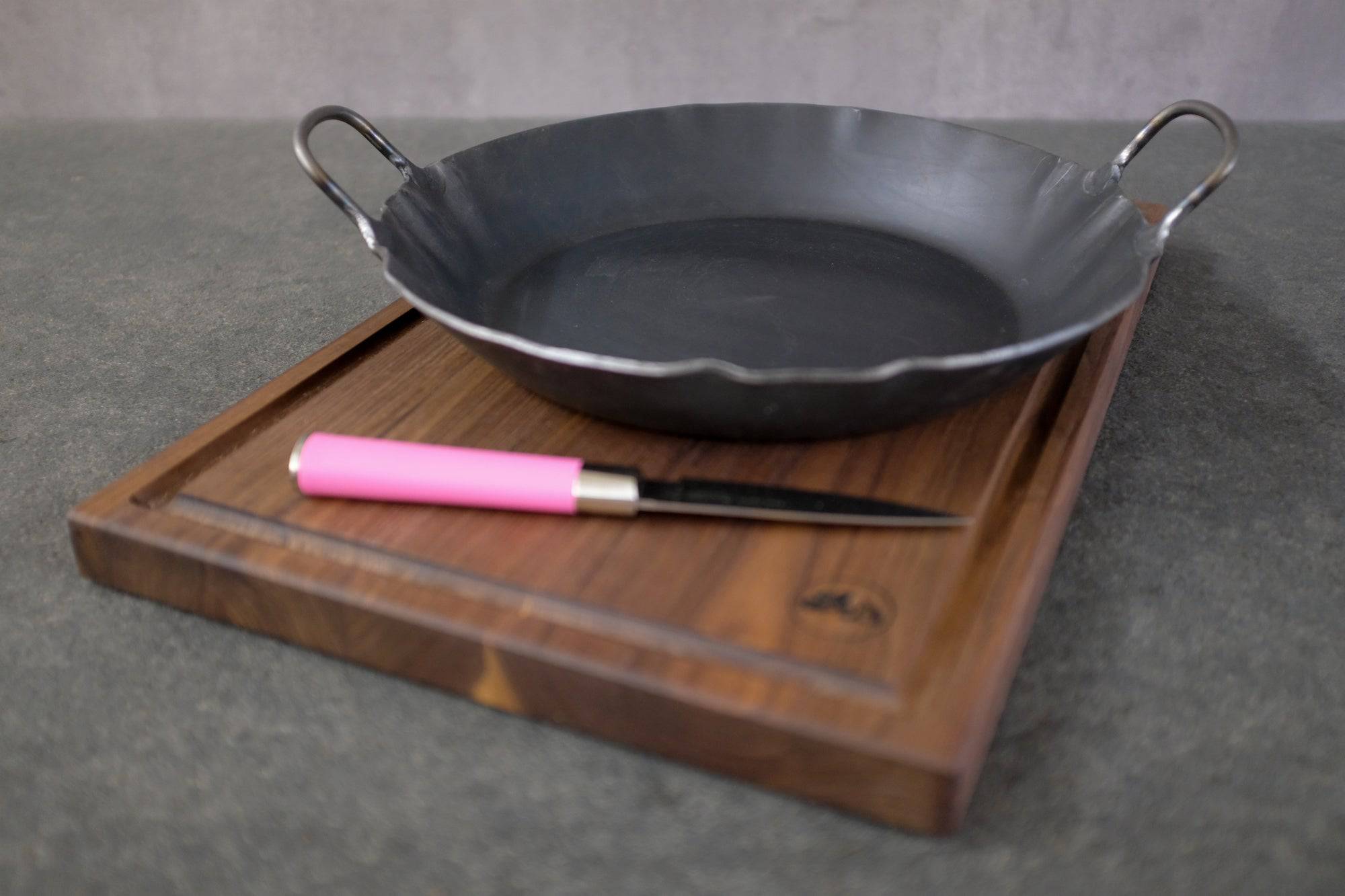 Servierpfanne aus Eisen mit zwei Griffen auf einem Saftrillenbrett aus Nussbaum und einem Dick Officemesser mit Griff in pink.