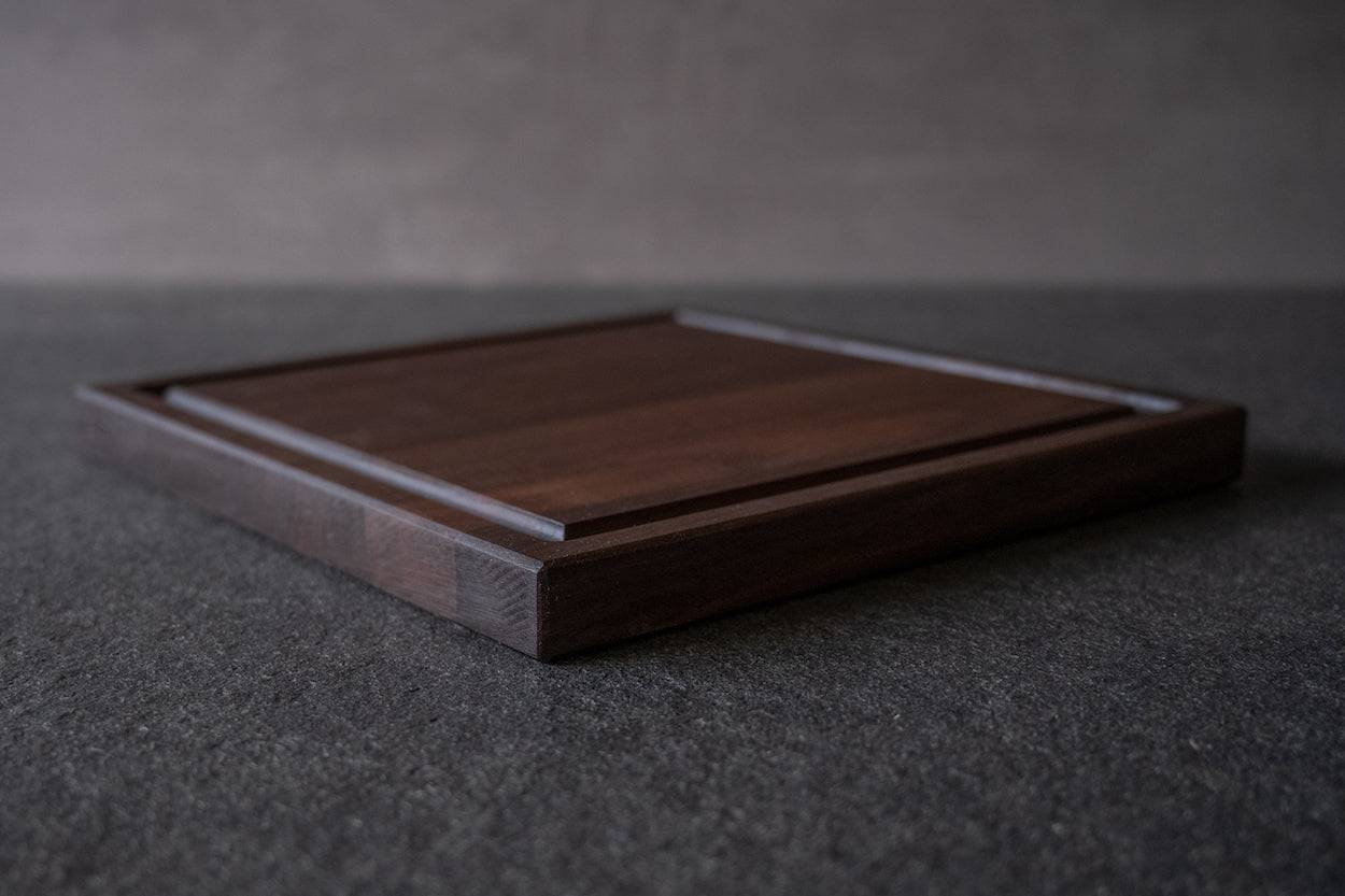 Saftrillenbrett, quadratisch, in Nussbaum auf einer Steinplatte liegend.