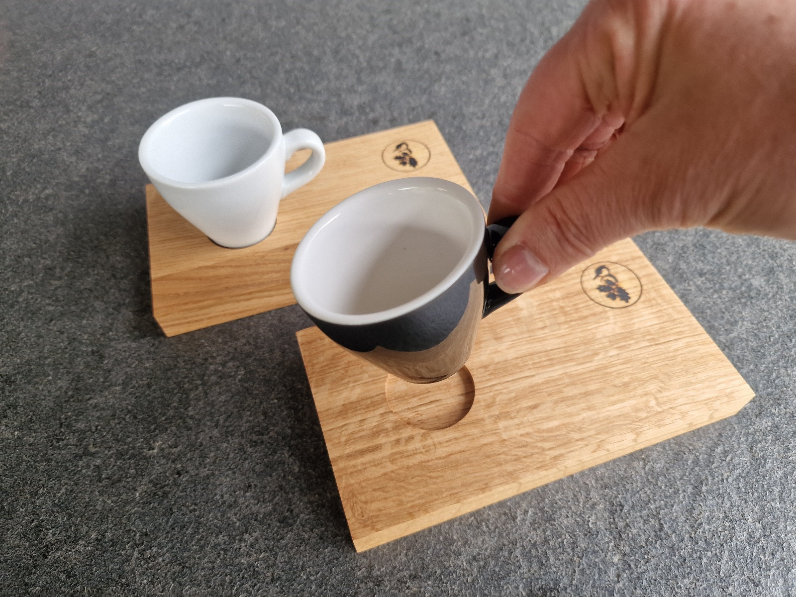 Eine Hand nimmt eine dickwandige Espressotasse von einem Holz Espressotablett. Im Hintergrund dasselbe mit einer weißen Tasse.