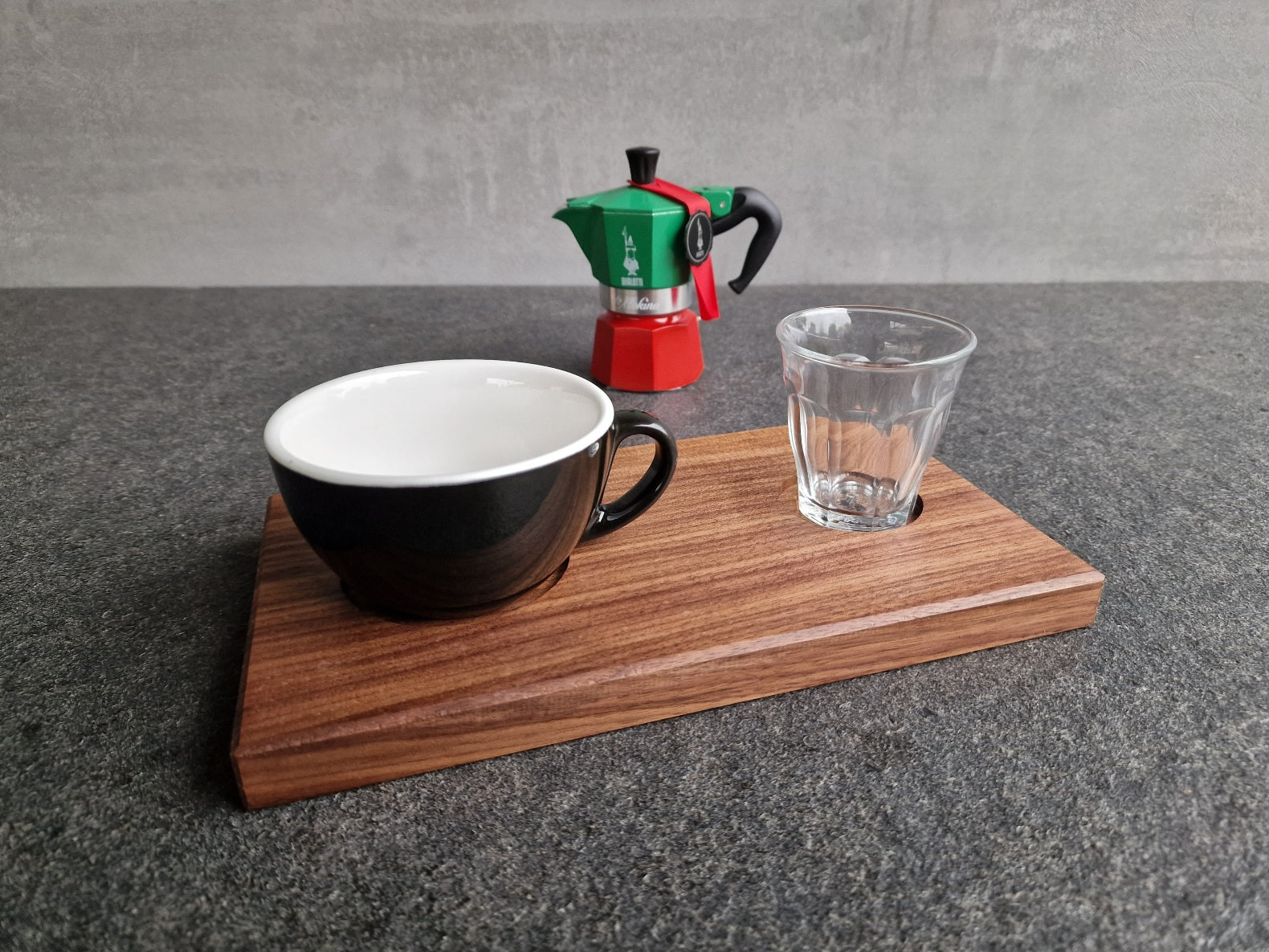 Nussbaum Tablett mit einer schwarzen Cappuccinotasse und einem Duralex Wasserglas. Im Hintergrund eine Bialetti Moka Espressokocher Tricolore.