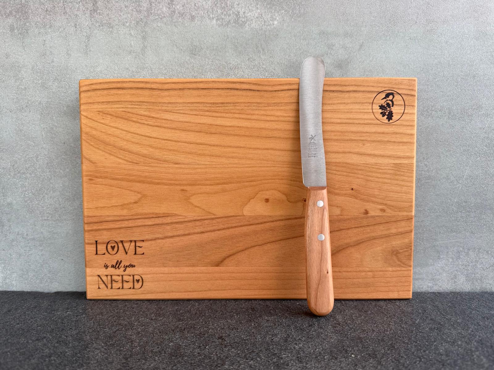 Kirschbaum Holzbrett mit Lasergravur für Valentinstag: Love is all you need. Dekoriert mit einem Brotzeitmesser mit Kirschbaumgriff von Windmühlenmesser.