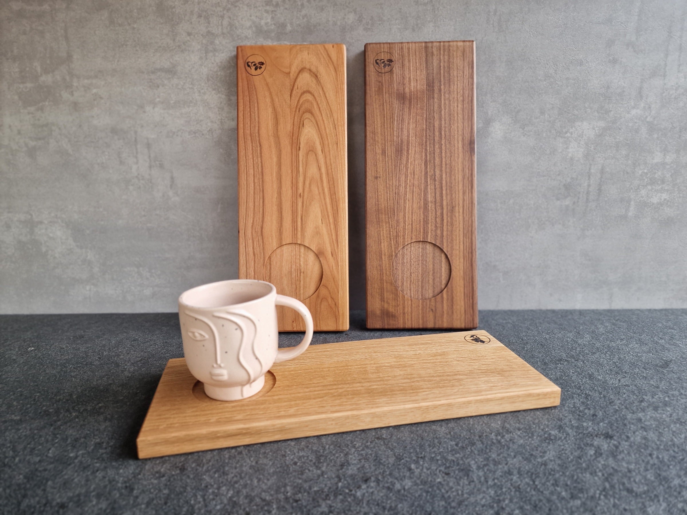 Cappuccino-Tablett aus Kirschbaum und Nussbaum an einer Wand angelehnt. Im Vordergrund ein Holztablett aus Eiche mit einer großen Tasse mit Gesicht.
