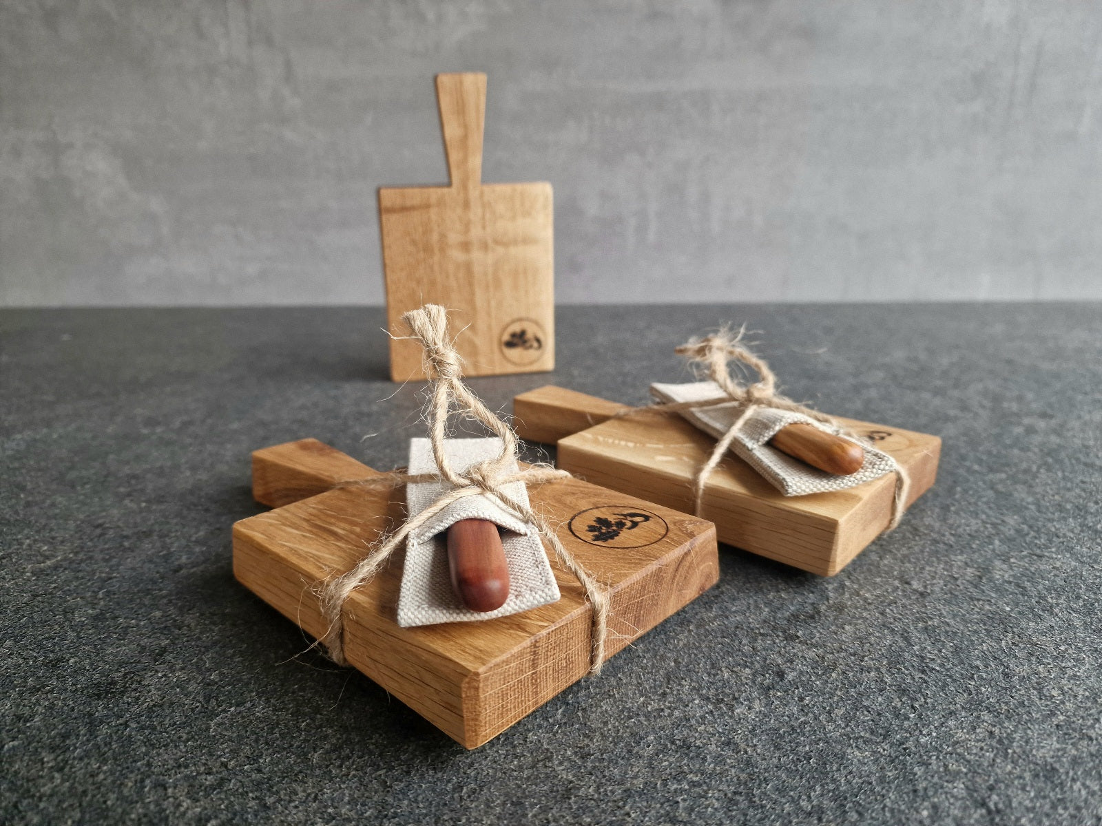 Ein Mini Eiche Holzbrett stehend im Hintergrund. Im Vordergrund zwei Holz Geschenke. Dasselbe Holzbrett jeweils mit einem Buttermesser im Etui verpackt, liegend auf einer Steinplatte.