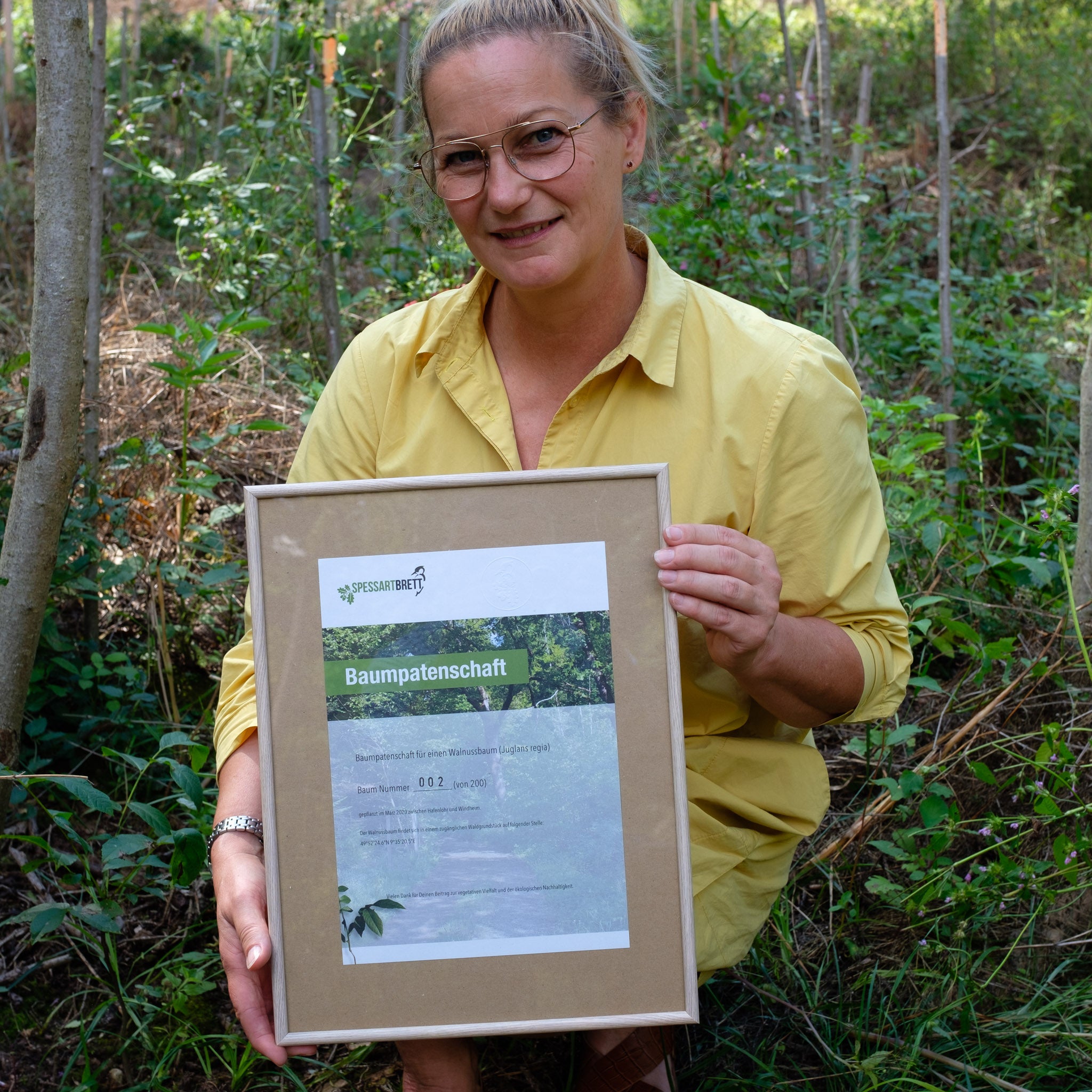 Eine Frau im Wald hält eine Baumpatenschaft Urkunde in einem Bilderrahmen in der Hand.